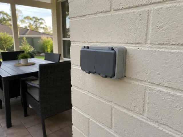 Outdoor double power point on a painted brick patio wall beside dining furniture in Holmesville